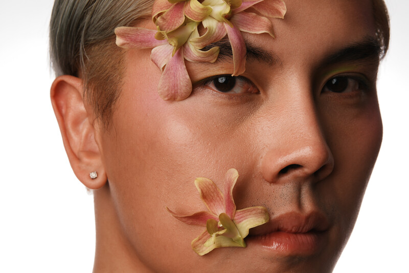 Close-up portrait of a person with wilted flowers positioned across their face against a white background.