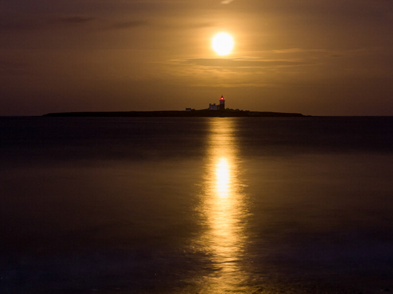 Golden sun reflecting across calm water toward a distant lighthouse at sunset.