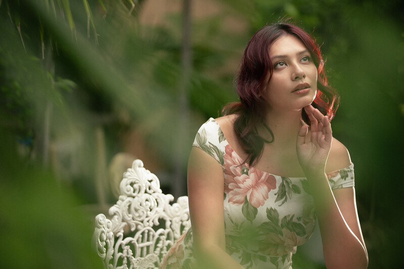 Woman in floral dress looking upward in soft-focus garden setting with white decorative element in foreground.
