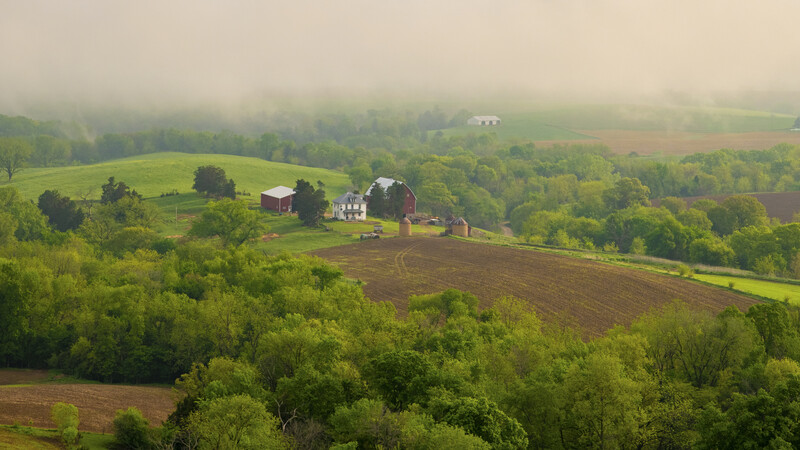 Aerial view of a farmhouse nestled among green fields and trees in rolling countryside.
