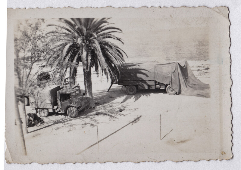Vintage black and white photograph showing a truck and tent camp beside a palm tree near water.