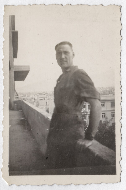 Man in dark jacket standing on a rooftop balcony overlooking a cityscape.