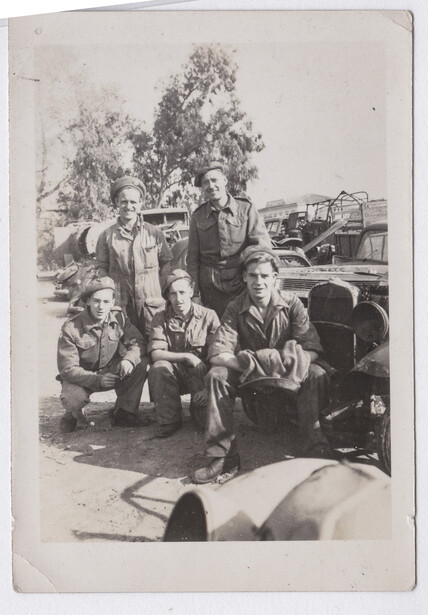Group of seven people posing together outdoors near vintage automobiles and trees.