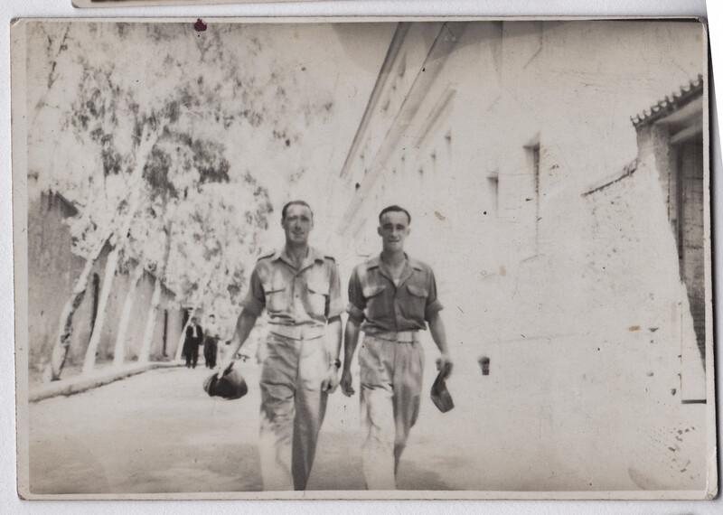 Two men in military uniforms walking down a sun-drenched street lined with white buildings.