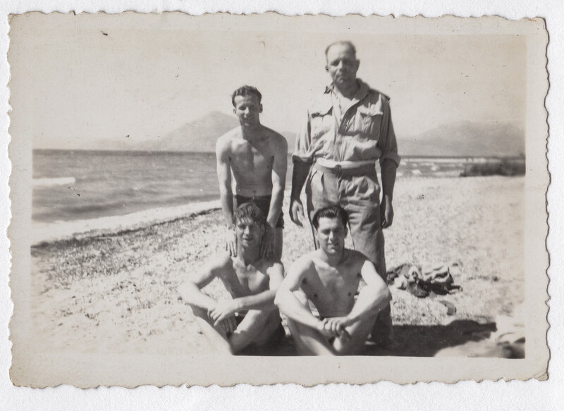 Four shirtless men on a pebbly beach, one standing in military uniform, three seated or crouching.