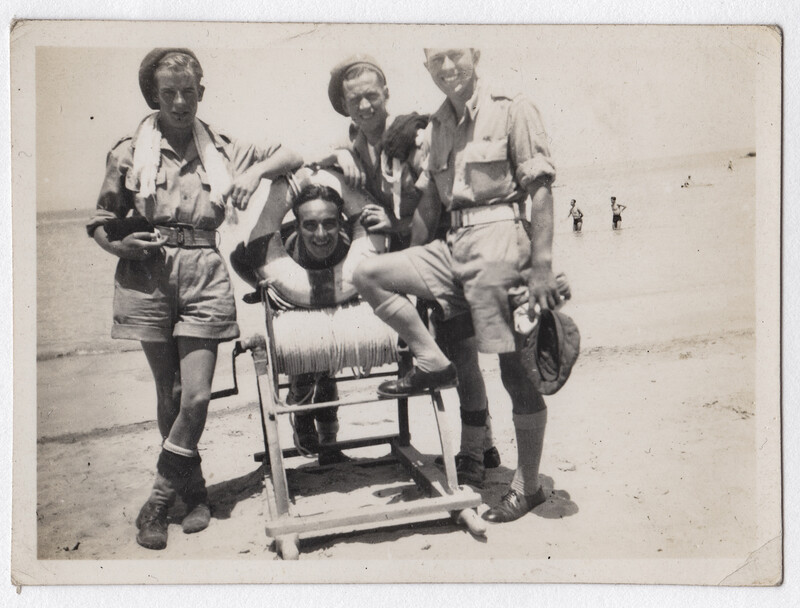 Four men in khaki uniforms posing with a vintage motion picture camera on a beach.