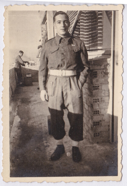 Man in military uniform standing indoors with boots and belt, vintage photograph.