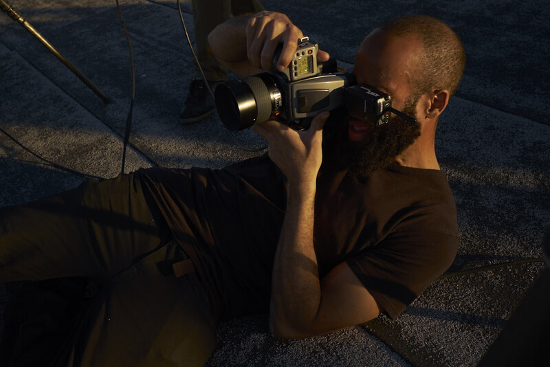 Photographer holding a camera with external flash, positioned low to the ground on a textured surface.