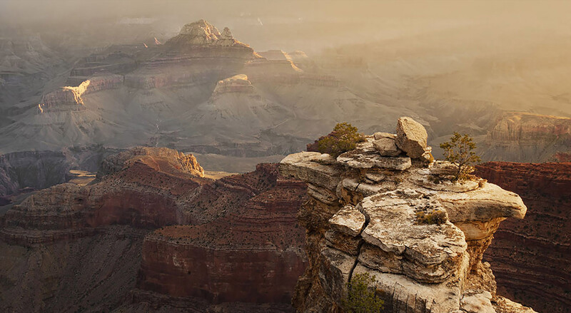 Rocky outcrop in foreground overlooking a vast canyon layered with stratified rock formations bathed in golden morning light.