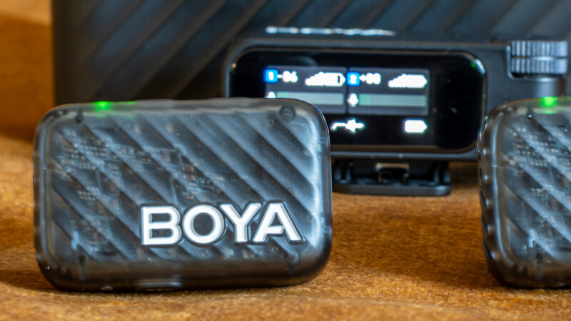 Two BOYA wireless microphone transmitters with textured black cases displayed on a wooden surface beside a receiver unit.