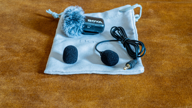 BOYA wireless microphone kit with windscreen, foam caps, cable, and carrying pouch on brown fabric background.