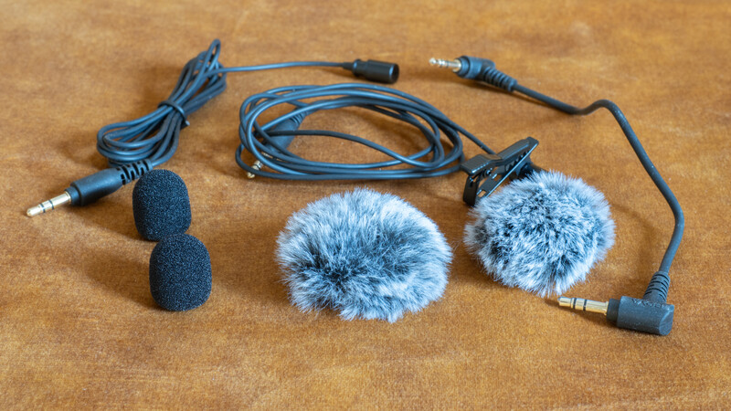 Lavalier microphone kit with foam and fur windscreens, extension cables, and connectors on tan fabric background.