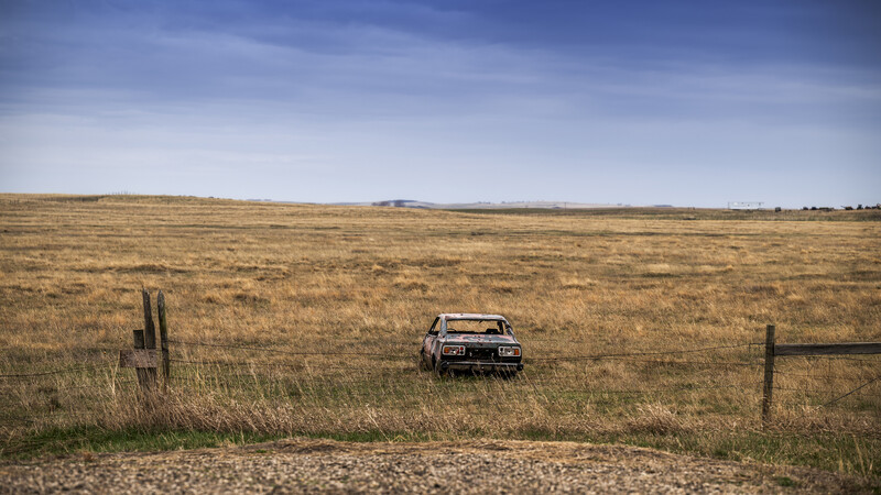 Abandoned vintage car sits alone in an expansive prairie field bordered by wooden fence posts.