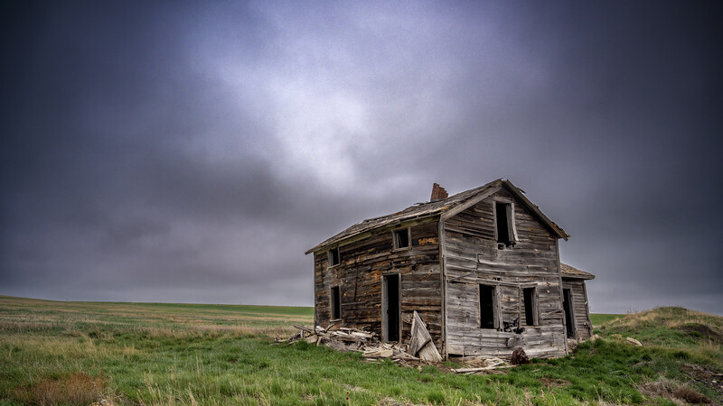Dilapidated wooden homestead standing alone in vast grassland under dramatic cloudy sky.