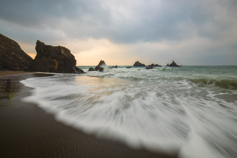 Waves crash onto a sandy beach with dark rocky outcrops scattered across the horizon under an overcast sky.