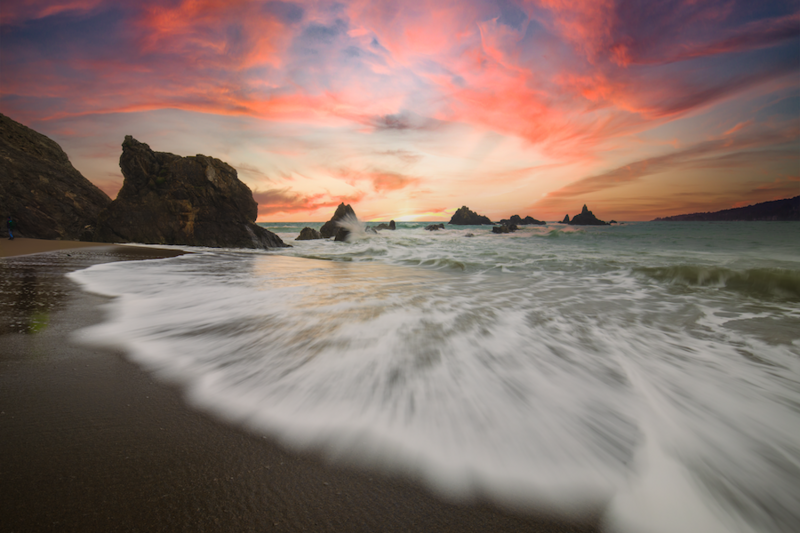 Waves wash across a sandy beach at sunset with rocky outcrops silhouetted against an orange and pink sky.
