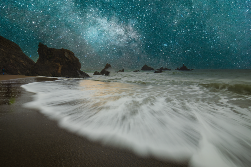 Long-exposure photograph of waves on a rocky beach beneath a starry night sky with visible Milky Way.