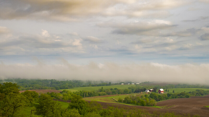 Rolling agricultural landscape with green fields, scattered trees, and farm buildings under partly cloudy sky.