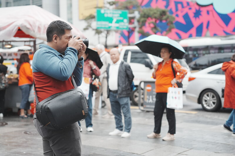 Photographer holding camera to face while shooting at outdoor event with colorful signage and people in background.