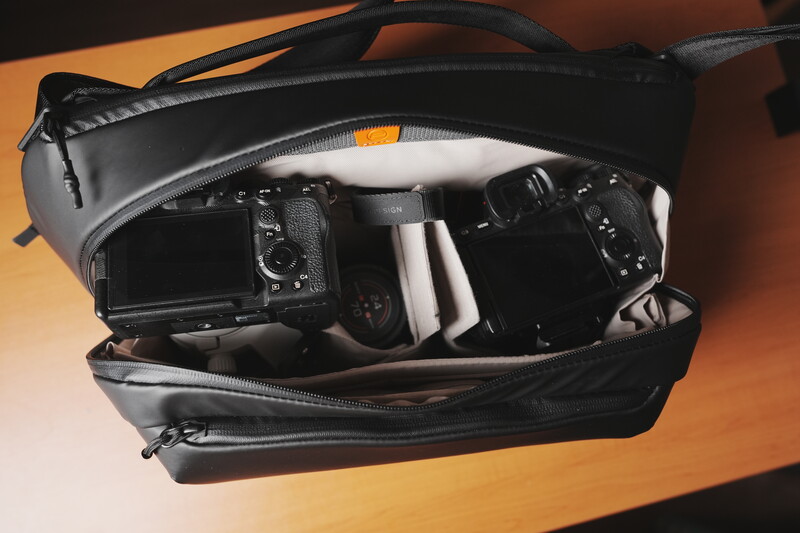 Black camera bag open on orange surface, revealing organized interior compartments with DSLR camera and lenses.