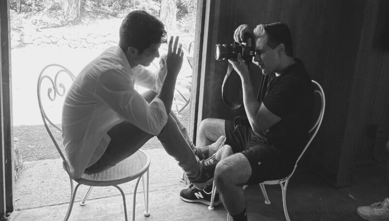 Photographer with camera photographing a seated subject posing by a window in black and white.