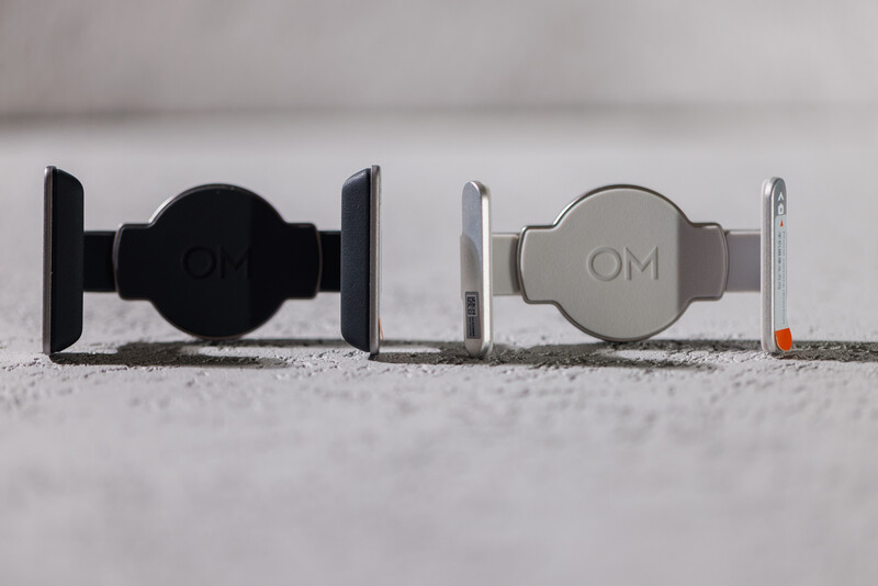 Two smartphone car mounts displayed side by side on a textured surface—one black and one silver with OM branding.