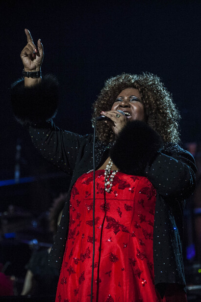 Performer on stage wearing a red patterned jacket, singing into a microphone with one arm raised.