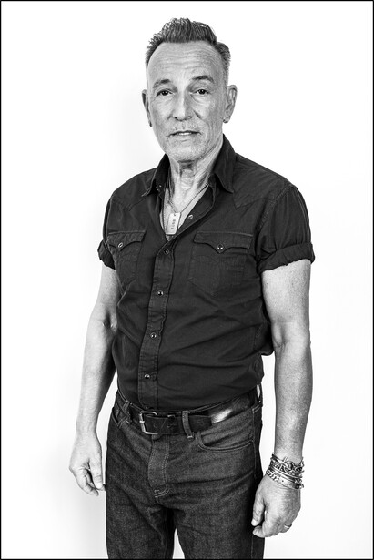 Black and white portrait of an older man wearing a dark short-sleeved button-up shirt and jeans against a white background.