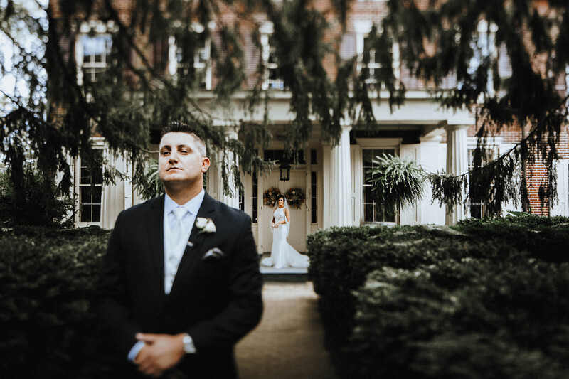Groom in dark suit standing in formal garden with white columned mansion in background.