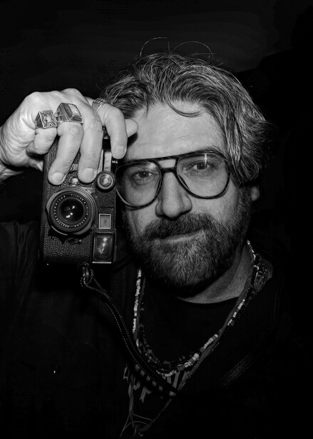 Bearded photographer wearing glasses holds a vintage film camera up to his face in a dark studio setting.