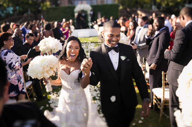 Bride and groom walk down outdoor wedding aisle with raised joined hands, smiling broadly amid guests and white floral arrangements.