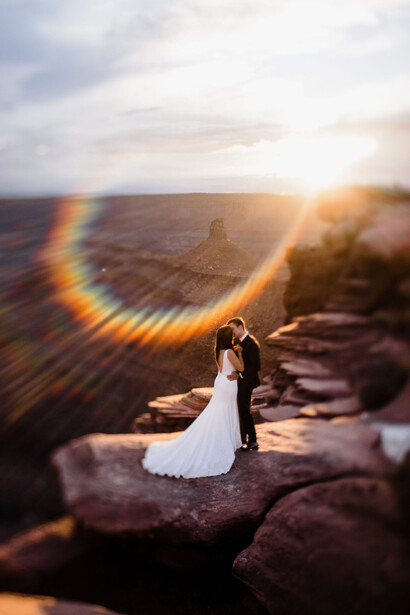 Bride in white dress standing on rocky outcrop at sunset, creating a dramatic rainbow light effect.