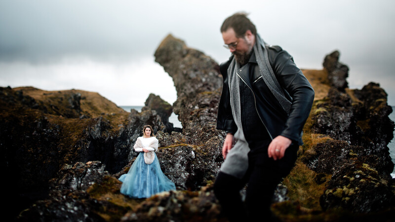 Man in dark jacket walking on moss-covered rocks toward a woman in a blue dress amid rocky coastal formations.