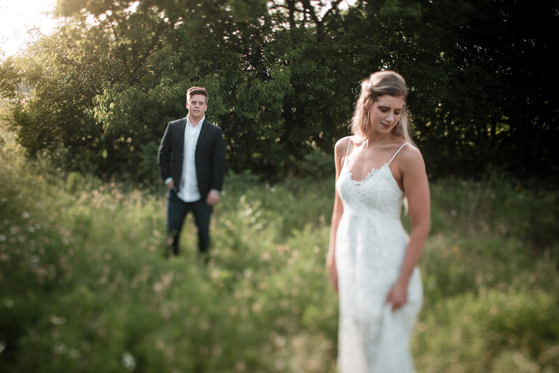 Bride in white dress smiling at camera with groom in dark suit standing blurred in background among green foliage.
