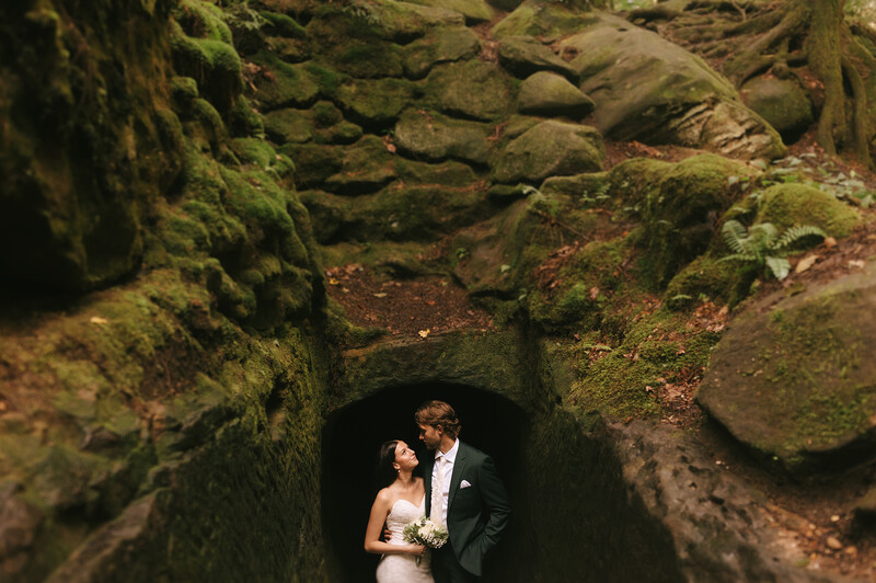 Bride and groom standing together in a narrow stone tunnel surrounded by moss-covered walls.