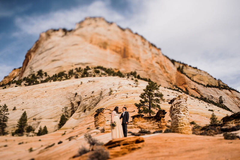 Couple standing together on desert terrain beneath a massive striated sandstone formation.