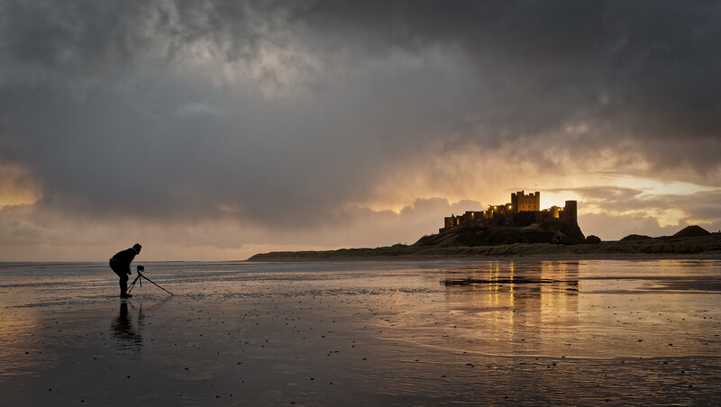 Photographer with tripod wading in shallow water at low tide, with a castle-topped island in dramatic backlighting.