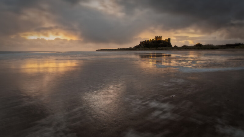 Silhouetted castle on a tidal island reflected in wet sand at sunset beneath dramatic clouds.