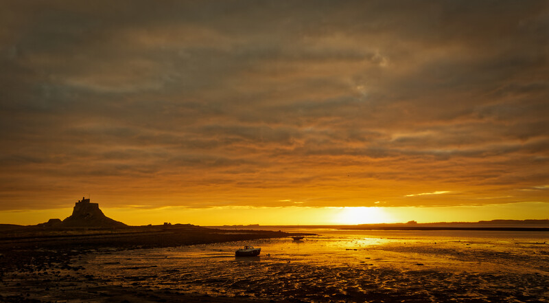 Golden sunset over a tidal bay with a silhouetted rocky island and a small boat on the exposed sand.