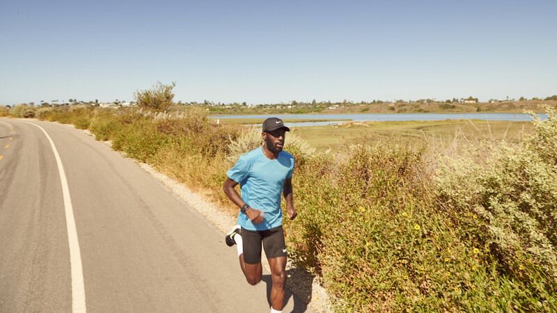 Male runner in blue shirt jogging along rural paved road with vegetation and wetland area visible in background.