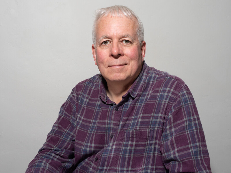 Older man with gray hair wearing a purple and blue plaid button-up shirt against a neutral gray background.