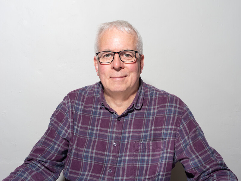 Older man with white hair and glasses wearing a purple plaid button-up shirt against a neutral gray background.