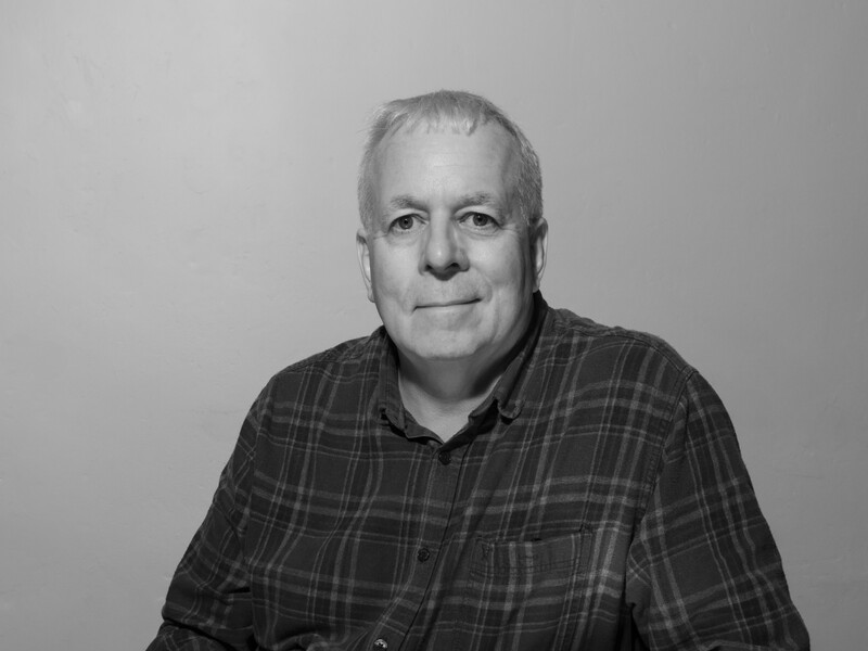 Black and white portrait of a middle-aged man wearing a plaid button-up shirt against a neutral background.
