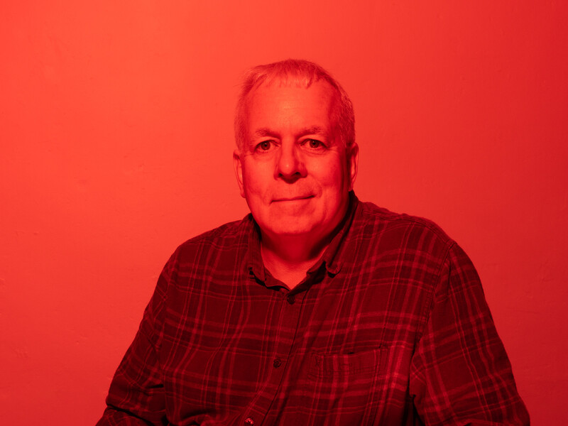 Middle-aged man wearing a dark red plaid shirt against a monochromatic red background.