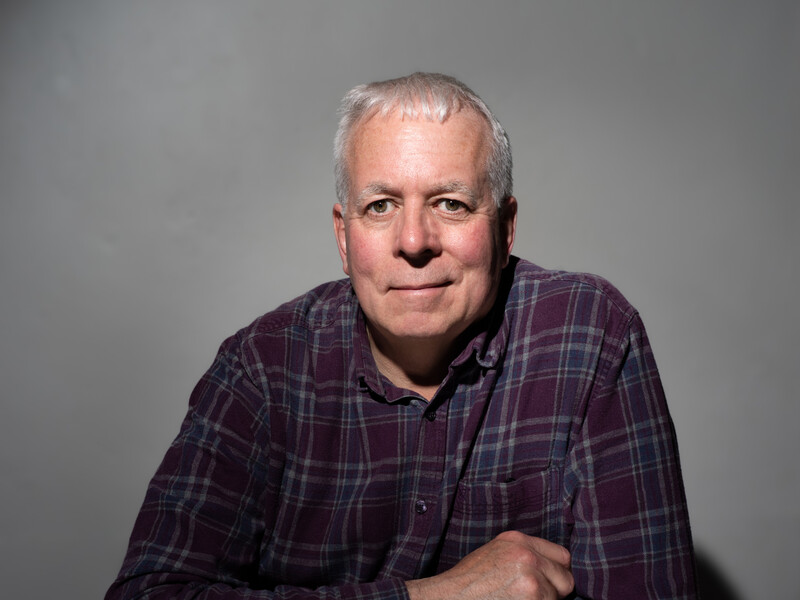 Middle-aged man in purple plaid shirt against neutral gray background.