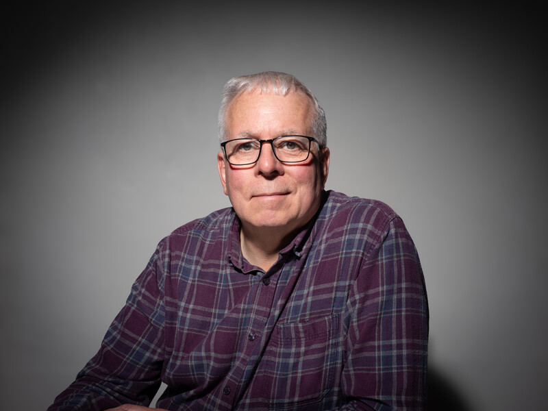 Middle-aged man with gray hair and glasses wearing a purple plaid shirt against a gray background.