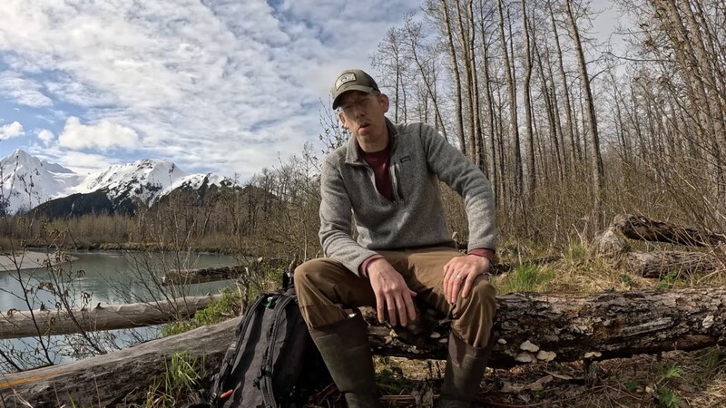 Man sitting on a log beside a river with snow-capped mountains and bare trees in the background.
