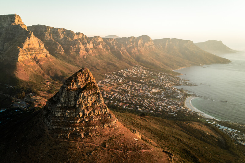 Dramatic rocky peak in foreground overlooking a coastal city nestled between mountains and sea at golden hour.