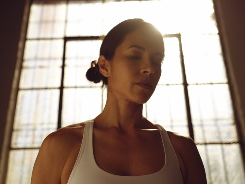 Woman in white tank top backlit by window with geometric panes, looking downward.