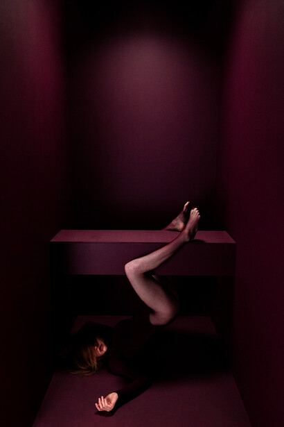 Nude figure in contorted position on wooden platform against deep burgundy background.
